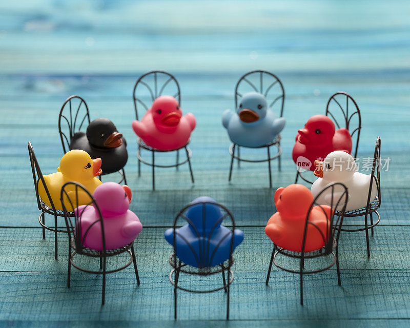 9只不同颜色的橡皮鸭坐在椅子上围成一圈，仿佛在开会，互相看着，设置在一个绿松石色的木头背景上。