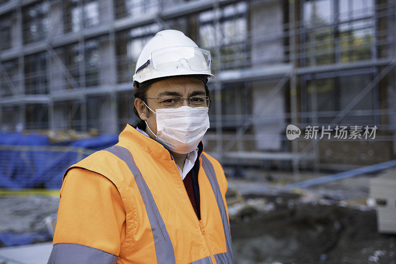 戴着防护服眼镜的男工程师微笑地站在正在施工的建筑前的工地上