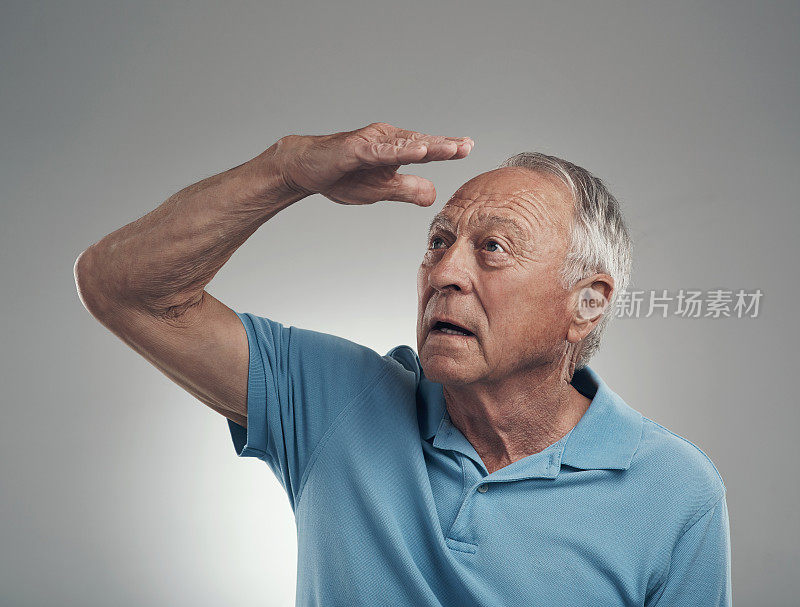 一个年长的男人用手捂住脸，在一个摄影棚里看着远方，以灰色的背景为背景