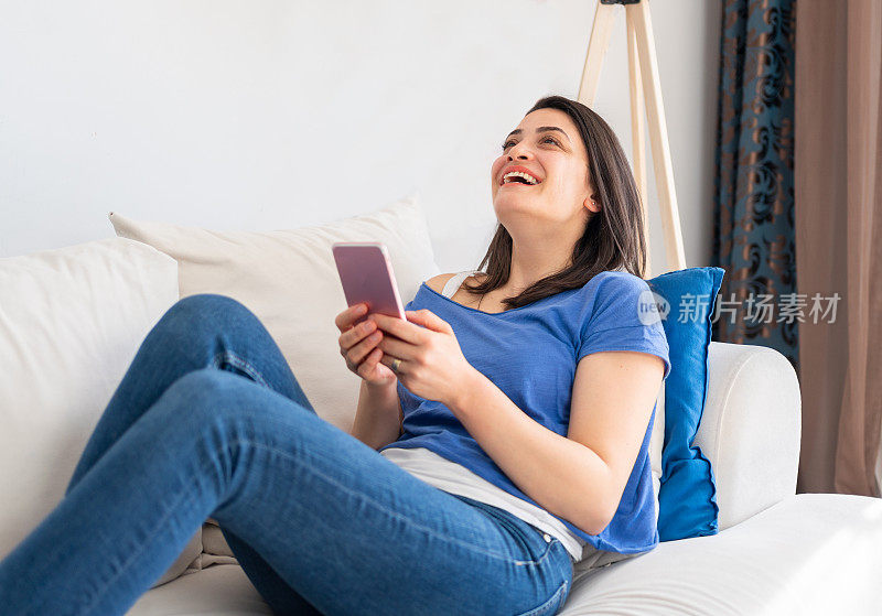 快乐微笑的女人躺在家里的沙发上使用智能手机。在家放松