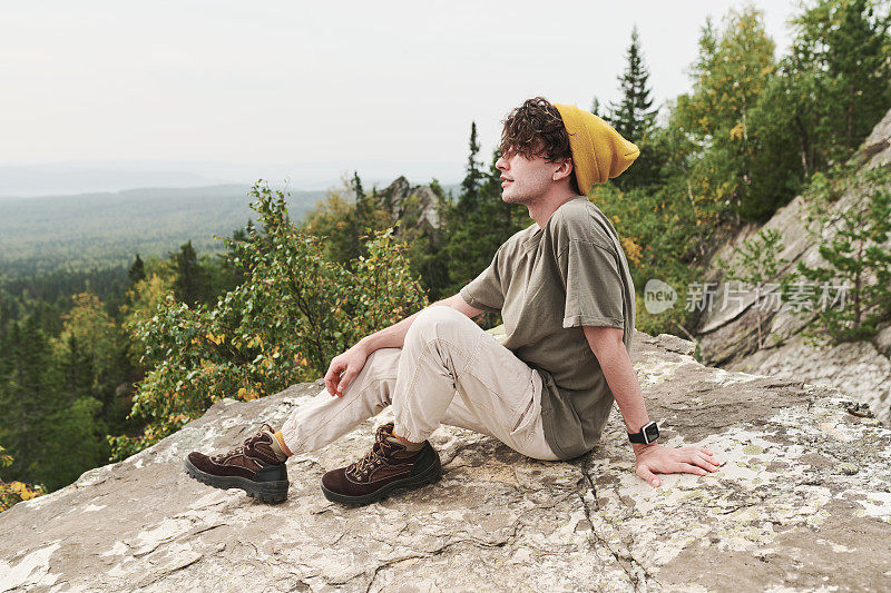 身穿黄色帽子和靴子的年轻高加索徒步旅行者坐在大石头上，凝视着森林景观