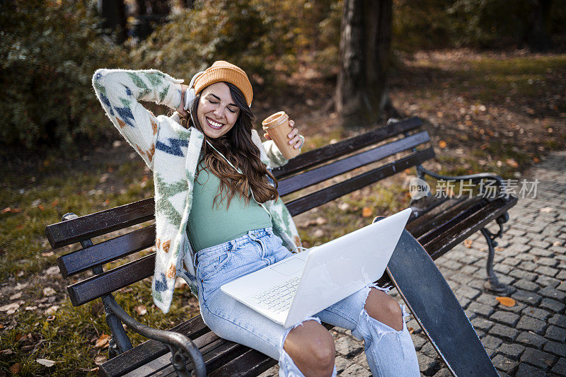 秋天阳光明媚的一天，一位年轻女子坐在公园的长凳上，用笔记本电脑和耳机听音乐