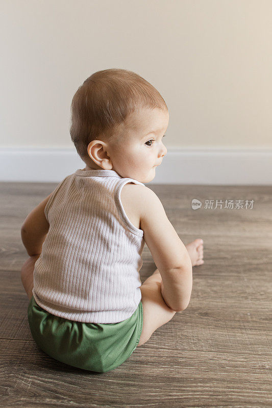 2022年，患有多指畸形的7个月大的男婴穿着橄榄绿可重复使用和可持续的布尿布和背心坐在家里的木地板上