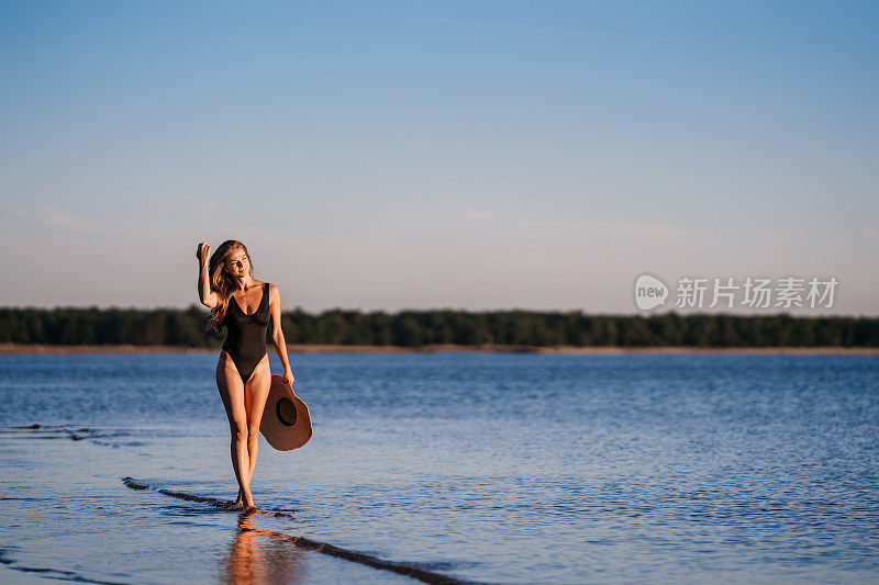 美丽的年轻健美女子在黑色泳衣走在海滩上，手持草帽在日落时分。成功的奢华女性度假时在岛上放松，享受海风。旅游及休闲活动