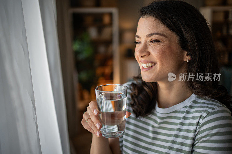 一个年轻女子站在窗边，用玻璃杯喝水。她的微笑反映了她关心自己的健康和适当摄入液体的意识。