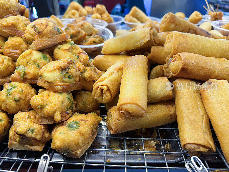 特写图片堆蔬菜春卷，面包(炸面饼)在金属格栅架与一次性，一次性塑料碗包含服务的部分在街头食品市场摊位上出售，不健康的街头食品外卖，重点在前景