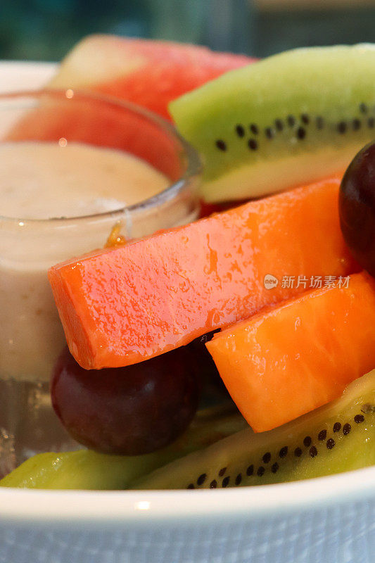 新鲜的全画幅图像，热带水果沙拉切片木瓜，菠萝，猕猴桃，西瓜，红葡萄在白色盘子周围的玻璃框架酸奶，重点在前景