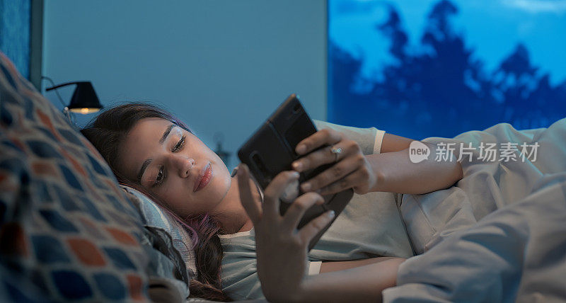 印度z世代伤心难过的女人躺在床上用手机聊天，深夜在家里的室内。孤独不快乐的女性在屋里看着智能手机屏幕打字读坏消息