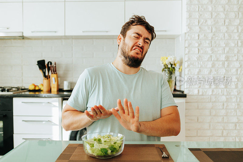 一个漂亮的年轻人坐在厨房里一碗沙拉前，双手放在身前。