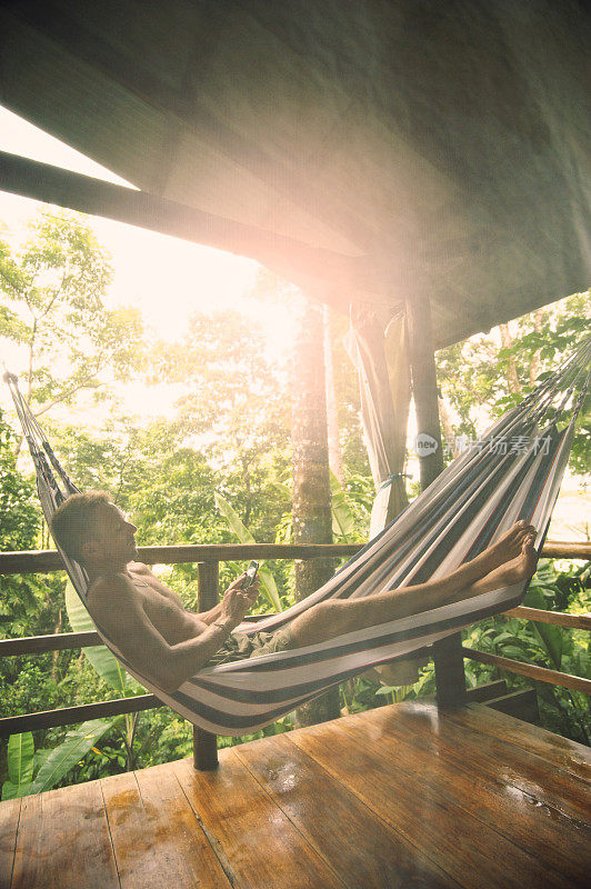 男子放松阳光丛林小屋吊床与智能手机蚊帐