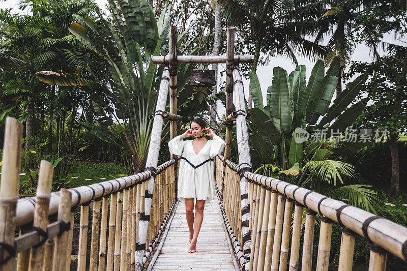 亚洲女孩在巴厘岛乌布的吊桥上行走。豪华别墅围绕苍谷绿色热带花园。