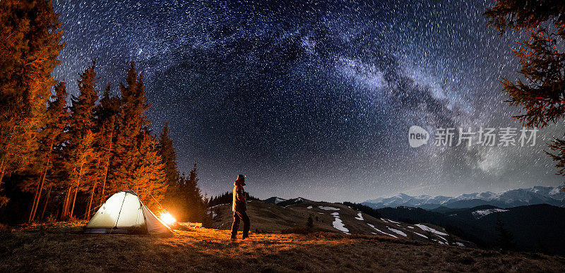 男游客晚上在森林附近的营地休息。一个人站在篝火和帐篷附近，在美丽的夜空下，满是星星和银河，欣赏夜景。全景景观