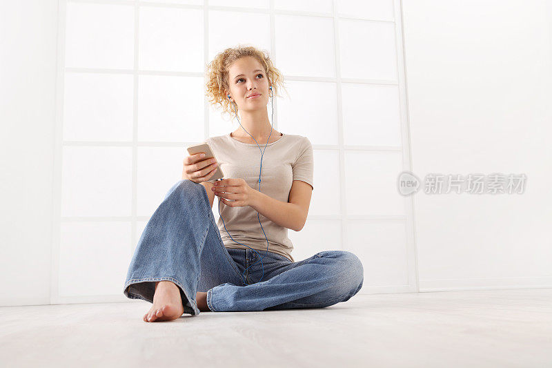 坐着的年轻女子，戴着耳机，拿着手机，卷着头发，穿着牛仔裤和T恤，地板上的白色房间背景被孤立
