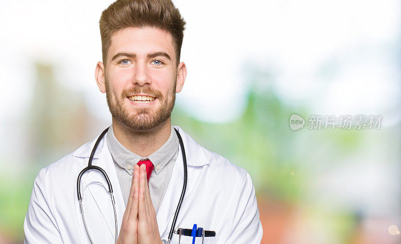 年轻英俊的医生男子穿着医衣祈祷与手一起请求原谅微笑自信。