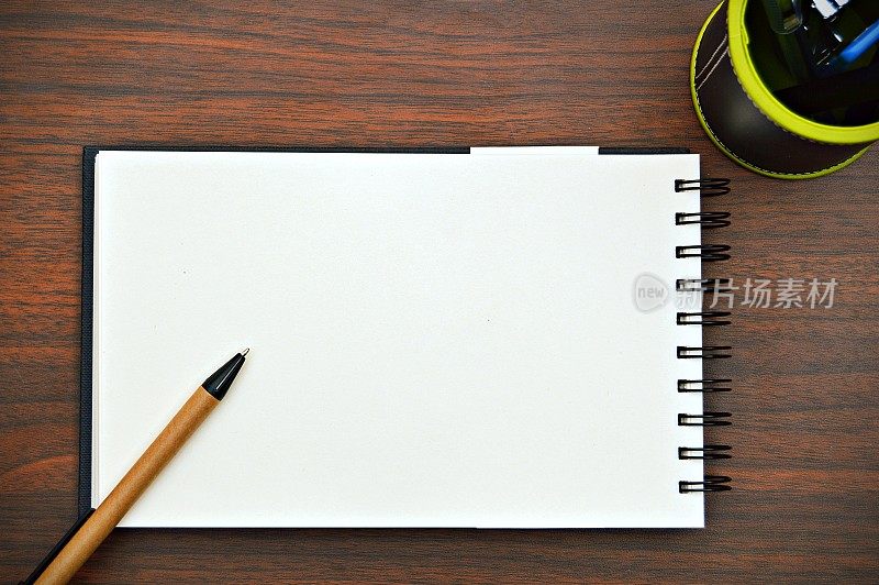 一个水平的照片，一支钢笔斜放在白色素色的螺旋式记事本上，放在一个木制的深棕色水平背景上，很有美感。