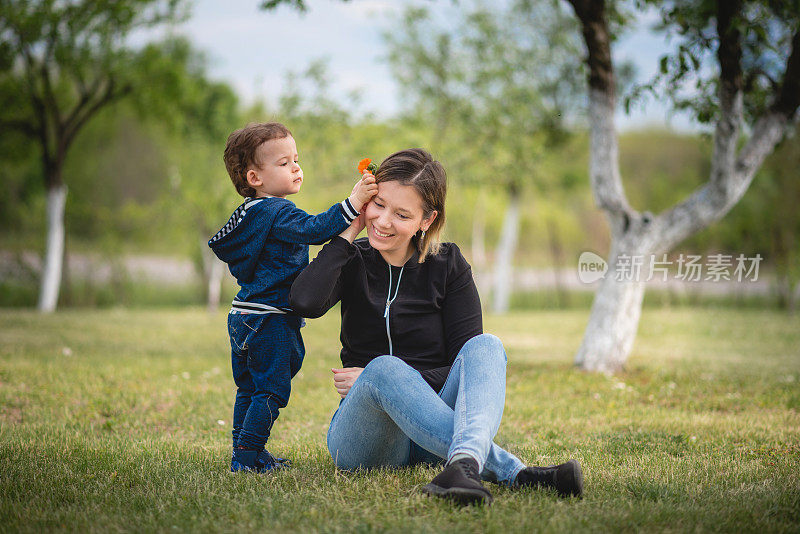年轻的母亲和她蹒跚学步的儿子一起庆祝母亲节，坐在后院的草地上享受他们的时光