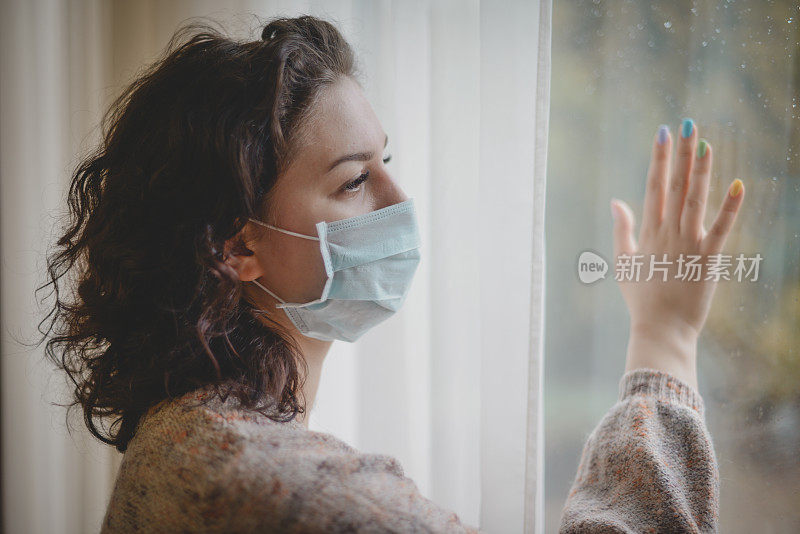 年轻女子戴着医用口罩，站在窗边