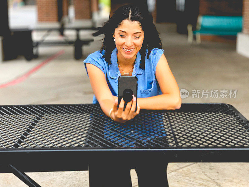 漂亮的混血女人坐在外面的桌子微笑和视频聊天与她的智能手机