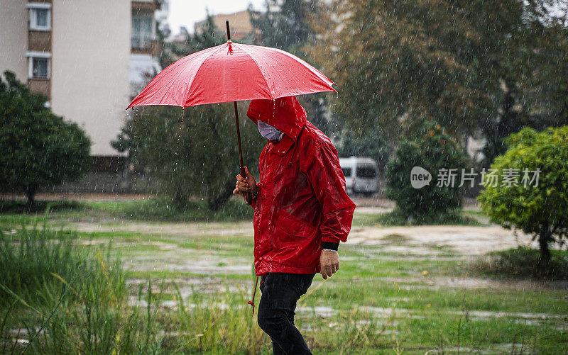 成熟的男人穿着红雨衣，撑着红伞走在雨中的城市街道上
