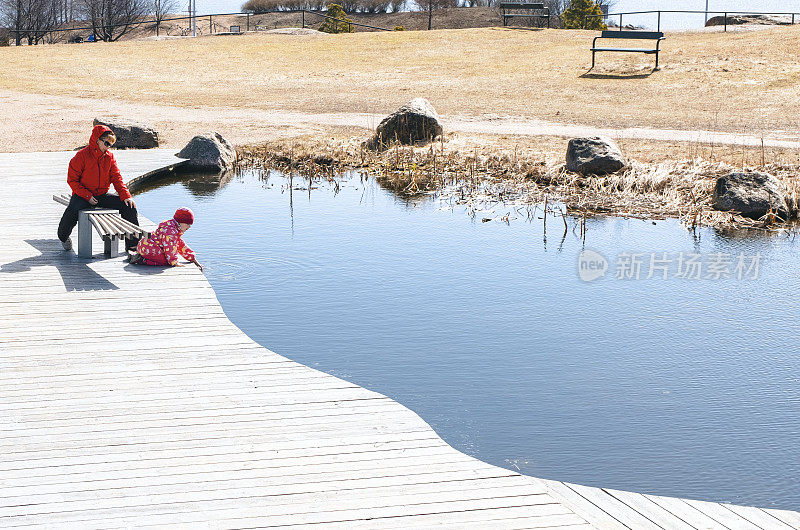一位妇女带着孩子在湖边的公园里散步。一个女人坐在公园的长椅上，一个小孩用手摸着水。湖上的木制装饰。一家白种人正在公园的大自然中休息。芬兰科特卡市凯瑟琳公园。