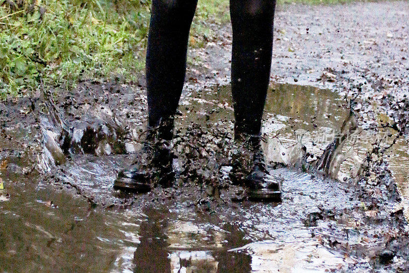 图像泥泞的乡村小路水坑与认不出的年轻女子划水，腿覆盖在黑色袜子和系带靴，同心涟漪在水面上，飞溅的水滴和水花，聚焦前景