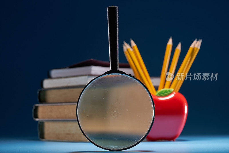 苹果形状的笔筒与一叠书和放大镜对蓝色的背景