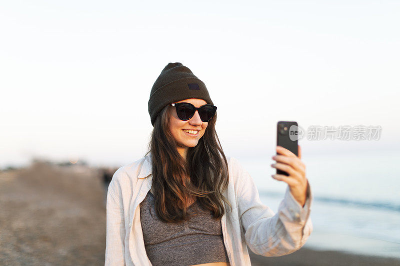 年轻的成年女子在日落时走在海滩上，使用手机