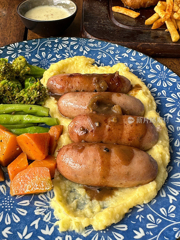 全帧图像的盘子传统的英国香肠和土豆泥，土豆泥顶部有两个猪肉香肠，绿色的西兰花，橙色的胡萝卜，覆盖在洋葱肉汁，丰盛的安慰食物，高处的视野