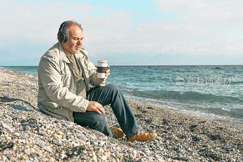 快乐的中年大胡子男子，戴着耳机听着智能手机应用程序的音乐或播客，坐在春天的海滩上喝着咖啡。放松的户外活动。