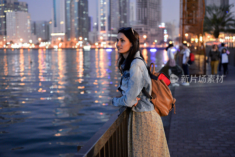 一个背着双肩包的游客女孩在夜晚欣赏着沙迦码头的现代摩天大楼