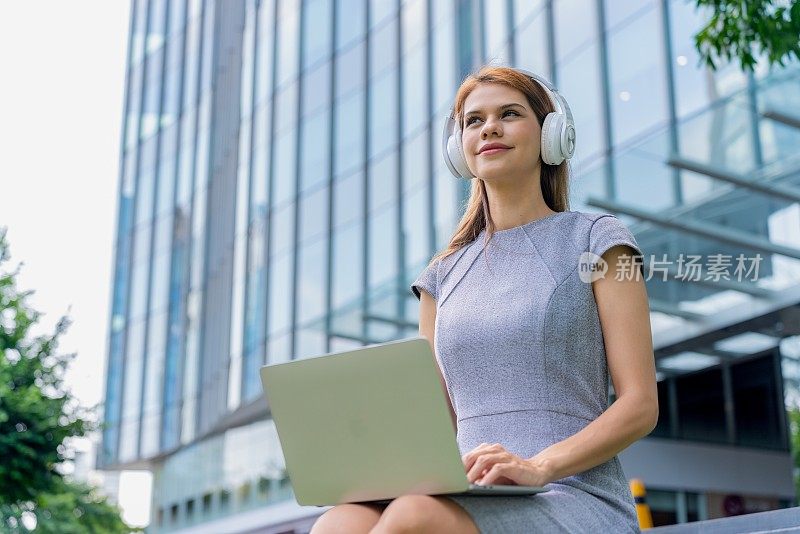 漂亮的女商人带着耳机和笔记本电脑在户外听音乐和看在线视频