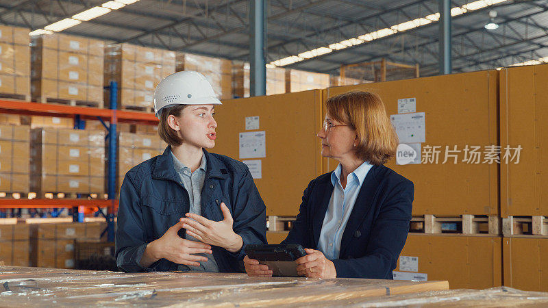 在零售仓库里，年轻的白人女工和成熟的女经理一起用数码平板电脑查库存并讨论谈话。物流行业业务。