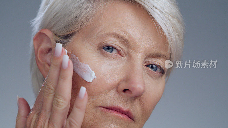 资深女性，在肖像上用面霜涂抹美容和护肤，在工作室背景上用手涂抹保湿霜。抗衰老化妆品护理，面部及女性特写用洗剂及皮肤病学