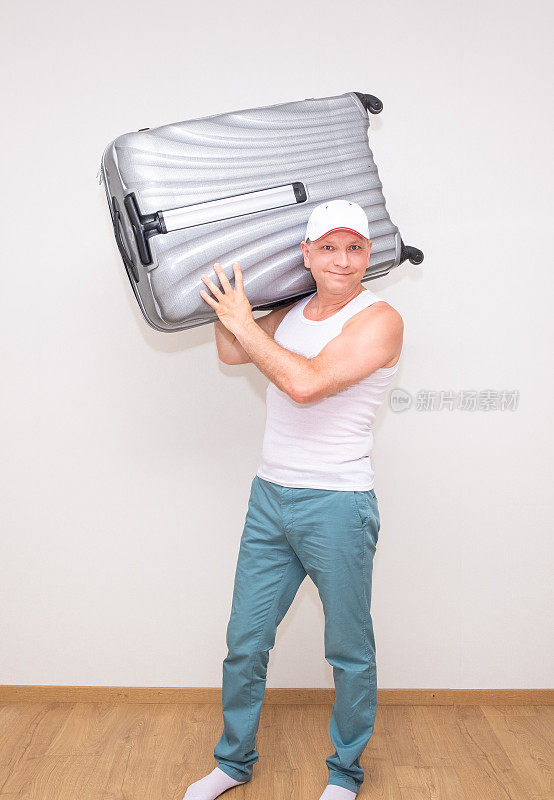 一个身穿白色t恤、头戴棒球帽的男人微笑着，肩上扛着一个大手提箱。准备出发。