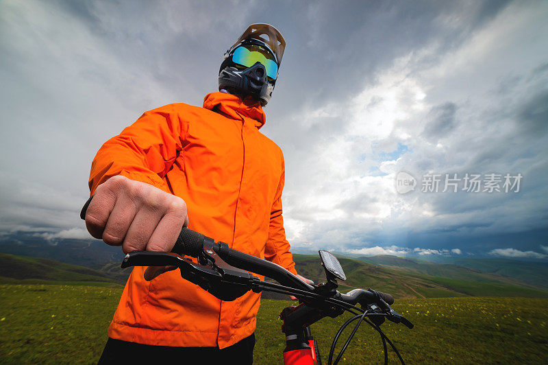 戴着全脸头盔和防护装备的骑手骑着一辆MTB自行车站在田野里，背景是乌云密布的群山