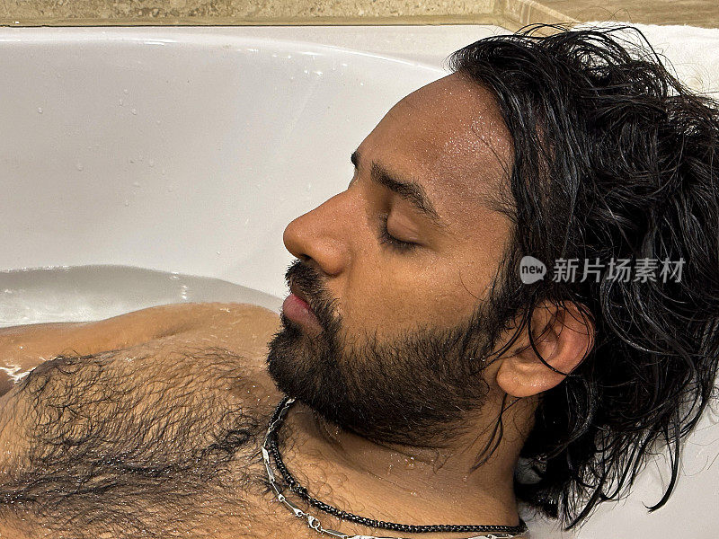 全画幅的印度男子躺在装满水的白色浴缸里，闭着眼睛，在浴缸里放松，头枕在浴盆边的毛巾上，毛茸茸的胸部，面部毛发，头像，侧面视图