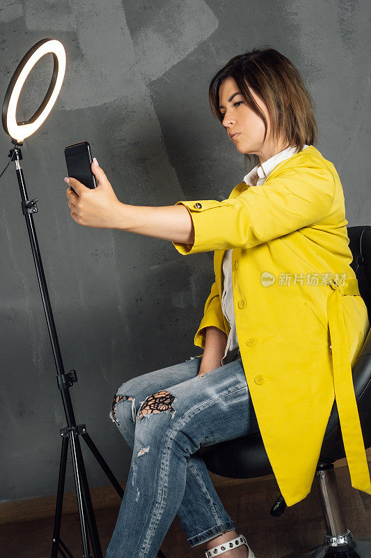 穿着黄色外套的时尚女士伸出手臂，拿着智能手机，坐在灰色的墙上自拍。