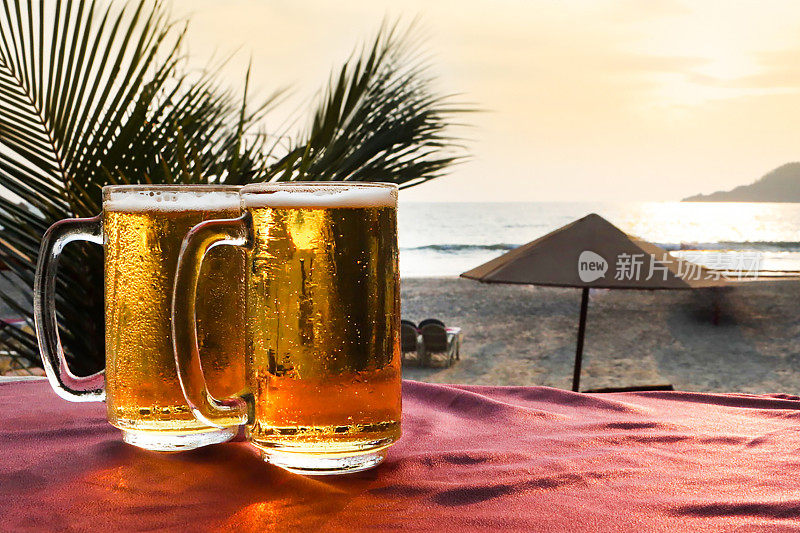 两个玻璃酒杯里的啤酒，上面满是凝结的气泡，在啤酒上形成泡沫的泡沫头，海滩酒吧的户外桌子上有桌布的马克杯，日落时沙滩上的遮阳伞，阳光明媚的橙色天空，重点是前景