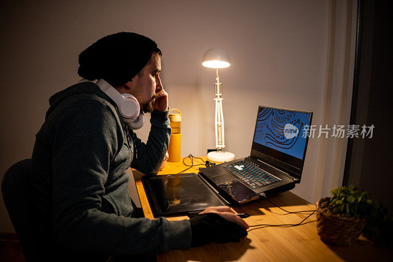 男性平面设计师在他的家庭办公室工作到很晚，同时使用笔记本电脑