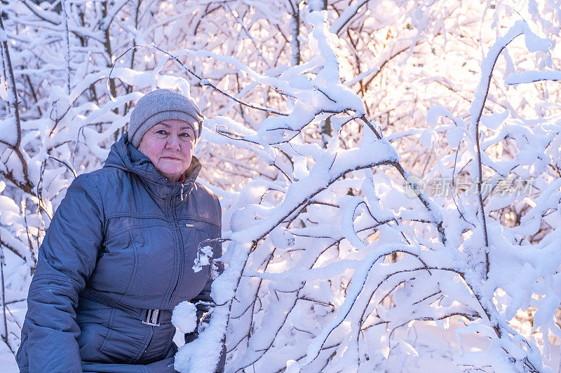 肖像。77岁的老妇人微笑着看着镜头，背景是白雪覆盖的树枝和阳光。