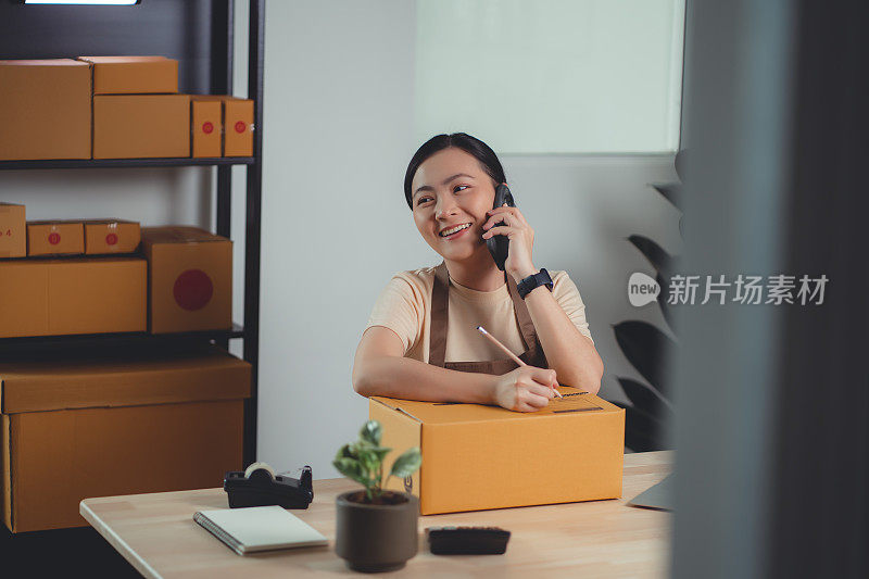亚洲女性在线卖家用智能手机打电话给客户确认订单，并包装产品准备发货。