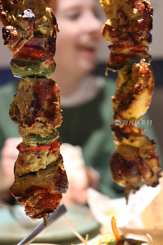 两张特写照片，挂在金属串架上的鸡肉串，烤焦的鸡胸肉块和覆盖着辣酱的红青椒串在一起，餐厅里的红发女人设置背景，重点放在前景上