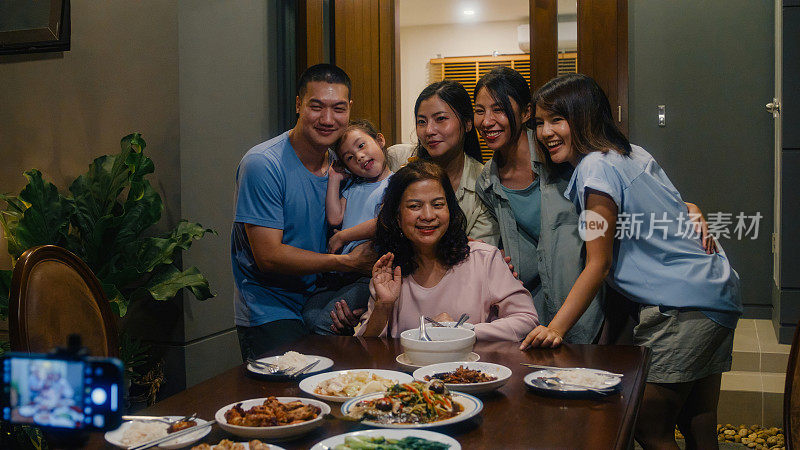 亚洲家庭用手机拍照，晚上坐在家外后院的餐桌上吃晚饭。多代同堂的家庭一起享受消费。