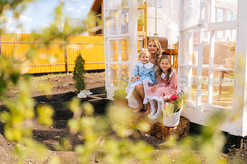 在阳光明媚的日子里，优雅的金发碧眼的年轻母亲和两个可爱的小女儿坐在夏日凉亭的台阶上