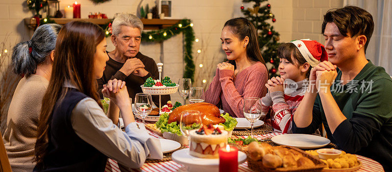 亚洲可爱的家庭在庆祝圣诞晚宴前祈祷。一群有吸引力的人在家里的餐桌上吃着庆祝节日感恩节和圣诞前夜的食物。