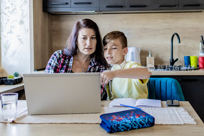 在家上学和做自由职业者。母亲在笔记本电脑上工作，而她的儿子在做作业或学习。他们在家里，坐在厨房的桌子旁，带着学习用品和笔记本电脑。