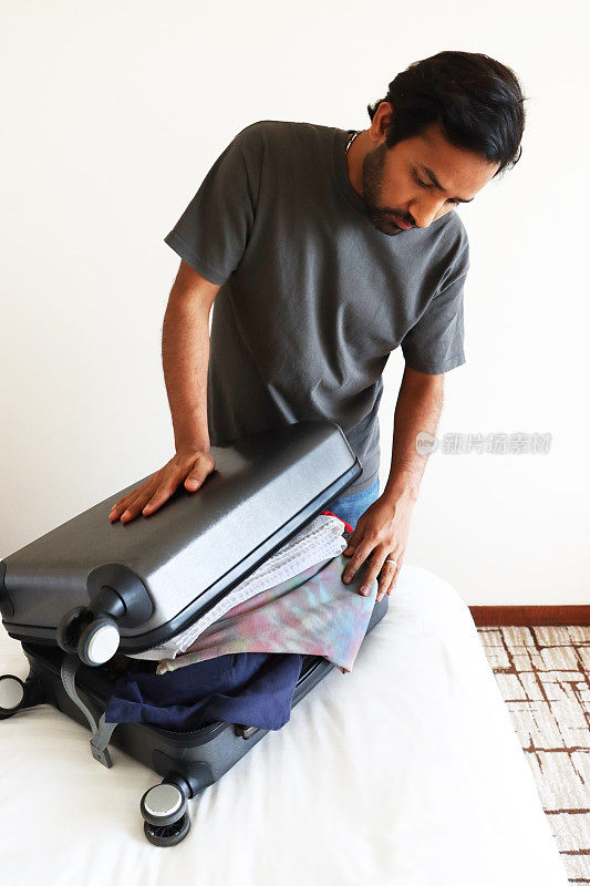 一名男子在铺着白色羽绒被的双人床上装着灰色的轮式手提箱，用手推着它盖上箱子盖，满是行李，整齐折叠的t恤从包里溢出，这是前景的焦点