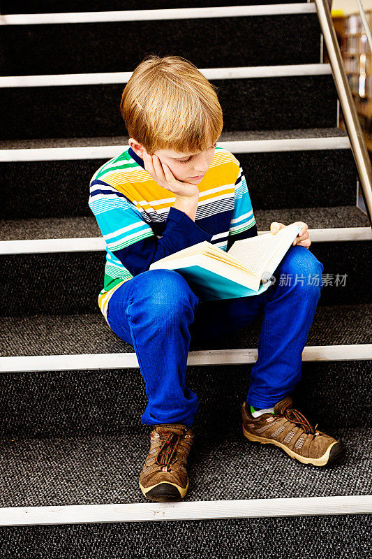 一个全神贯注的金发男孩坐在图书馆的台阶上看书