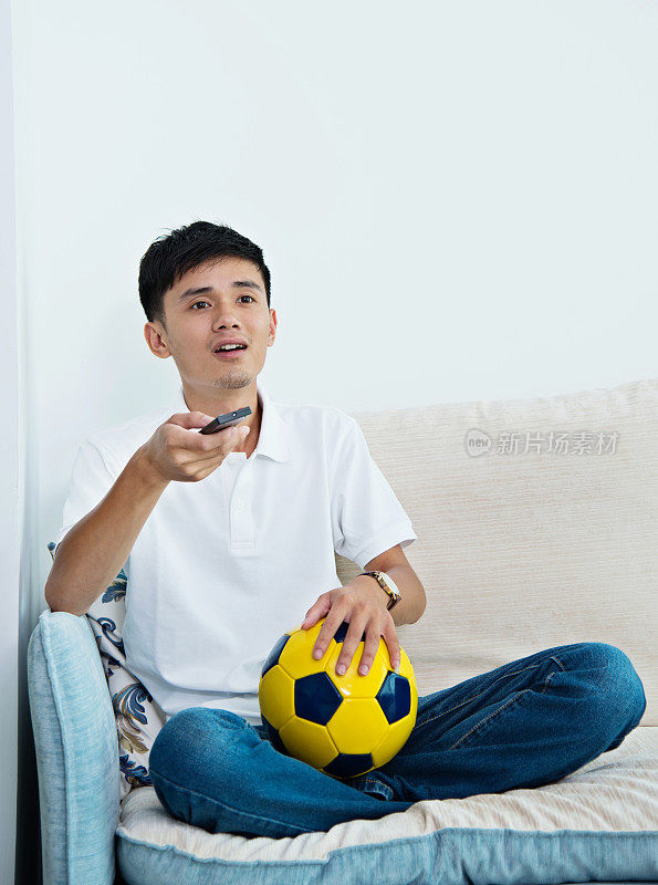 一个男人正在家里看电视，手里拿着足球