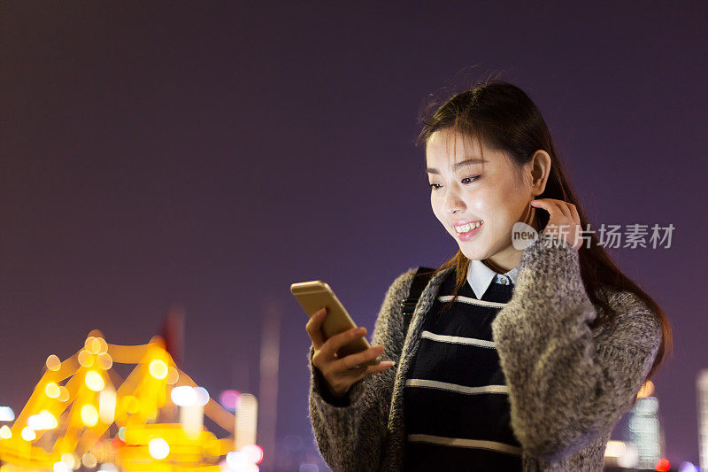 年轻美丽的亚洲女人晚上在现代城市玩手机